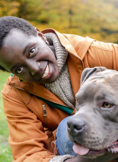 Close portrait smiling dark-skinned woman in orange coat hugging grey dog on autumn grass, emotional closeness, trust, calm mood during outdoor time in colorful seasonal park environment.